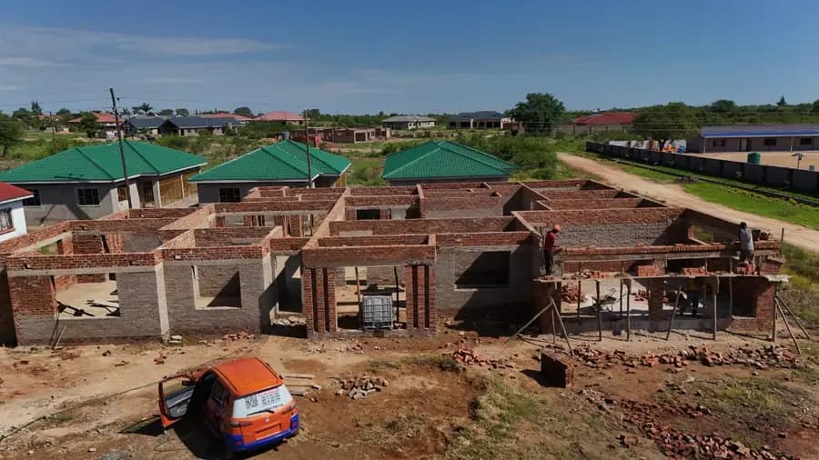 The brick shell of a large house under construction, walls up to lintel height, with finished green-roofed houses behind