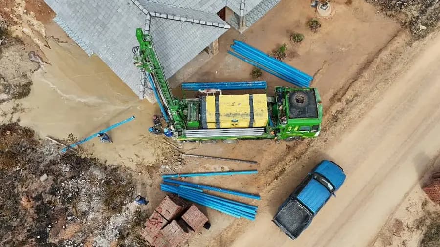 Aerial view of a green truck-mounted drilling rig on a stand, blue pipe and brick stacks laid out nearby