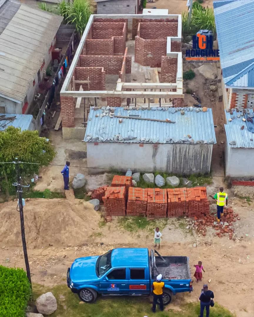 Bricklayers working on a house under construction beside a blue-roofed shed, with brick stacks and a bakkie on site