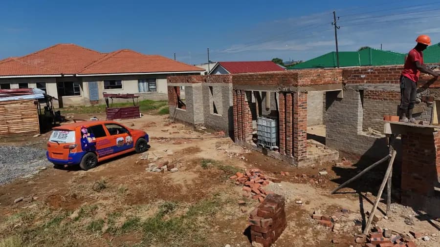 A brick house under construction with a red sheeted roof, a bakkie parked on the sand outside