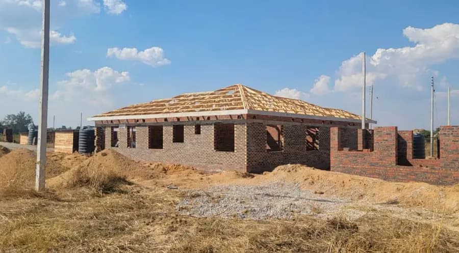 A timber hipped roof structure framed over the brick walls of a house on a dry, open plot